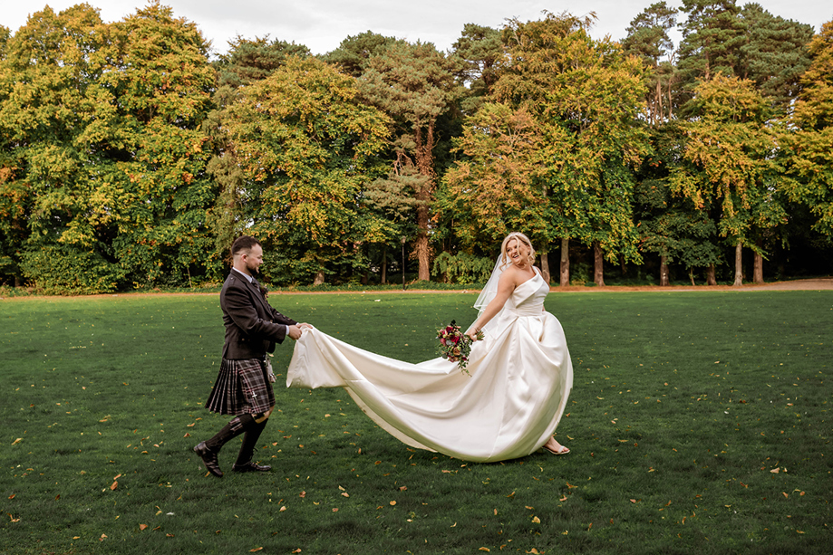 Groom carries his brides train as she runs across grass